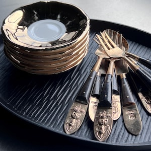 Peut inclure: Une pile de soucoupes noires à bordure dorée avec un centre blanc, et un ensemble de fourchettes, cuillères et couteaux dorés avec des manches noirs et des motifs ornés, disposés sur un plateau noir texturé.