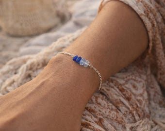 May include: A delicate silver bracelet featuring three blue and clear gemstone beads. The bracelet is worn on a wrist, with a soft, patterned fabric in the background. The gemstones are a focal point, adding a touch of color and elegance.