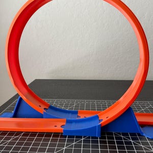 May include: A bright orange and blue toy race track loop. The loop is a complete circle, supported by blue track pieces. The track is designed for toy cars. The loop is made of plastic.