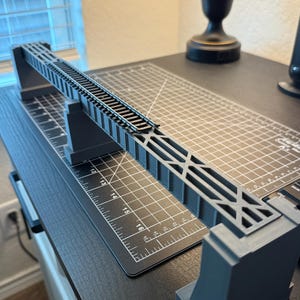 May include: A gray model train bridge with detailed track and support structure. The bridge is elevated and appears to be made of plastic or a similar material. The design includes cross-bracing and is set on a black surface.