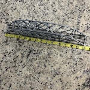 May include: A gray model bridge with a truss design, approximately 14 inches long, is shown with a yellow measuring tape. The bridge has a detailed structure with cross-bracing and is likely for a model train set or diorama.