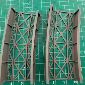 May include: Two gray, 3D-printed architectural models. Each model has a rectangular frame with an intricate, crisscross lattice design. The models are positioned on a green cutting mat with measurement markings.