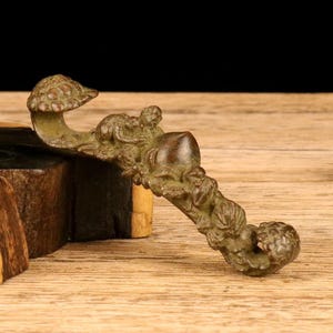 以下が含まれることがあります： キノコ型のボウルと、彫刻された猿などの姿が特徴のブロンズ色の装飾パイプ。木製の表面に置かれ、その精巧な職人技が際立っています。