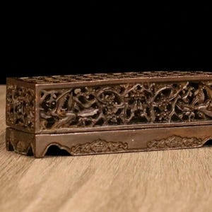 May include: A rectangular, brown, carved wooden box with intricate floral and bird designs. The box has a hinged lid and sits on a wooden surface. The carvings are detailed and create an openwork pattern.
