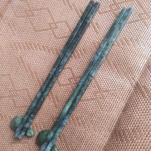 May include: Two jade green chopsticks with matching rests. The chopsticks are rectangular with a slightly textured surface. The rests are rounded and have a similar green hue. The chopsticks are angled across a textured, tan surface.