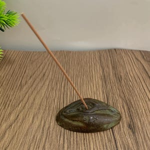 Può includere: Un porta incenso color bronzo con una forma organica strutturata. Un bastoncino di incenso è inserito nel supporto, inclinato verso l'alto. Il supporto ha una finitura screziata verde e marrone, che suggerisce un aspetto invecchiato. Lo sfondo è una superficie di legno.