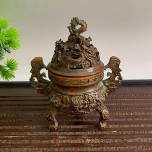 Può includere: Un bruciatore di incenso in bronzo con intricati dettagli di draghi. Il bruciatore presenta una base a tre gambe, manici ornati e un coperchio decorativo con una scultura di drago. Il design generale è dettagliato e invecchiato.