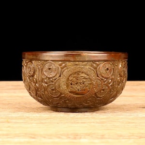 May include: A bronze-colored, intricately carved bowl. The bowl features detailed relief carvings of circular medallions and figures. The rim and base have a darker hue than the body of the bowl. The bowl sits on a wooden surface.