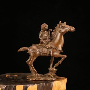 May include: A bronze sculpture of a figure riding a horse. The rider is seated on the horse, which is rearing up on its hind legs. The sculpture is set on a wooden base against a black background.