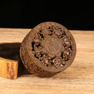 May include: A detailed, brown, round box with intricate carvings. The lid features a central design with Chinese characters, surrounded by floral and animal motifs. The box is made of a dark, possibly wooden material, and sits on a wooden surface.