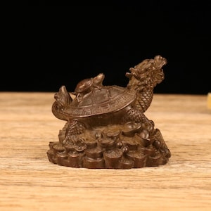 May include: A bronze-colored figurine depicting a turtle with a dragon's head and a smaller turtle on its back. The figurine sits atop a base of coins, showcasing intricate details and textures.