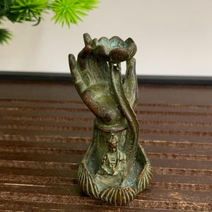 May include: A bronze-colored sculpture of a hand holding a lotus flower, with a small seated figure inside. The sculpture has a weathered, antique appearance, with green and brown tones. The hand is detailed, and the lotus flower is open.