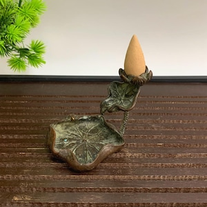 May include: A bronze incense holder featuring a lotus leaf design. It has a small dish and a leaf-shaped support for the incense cone. The incense cone is light brown and cone-shaped. The holder is on a dark brown surface.