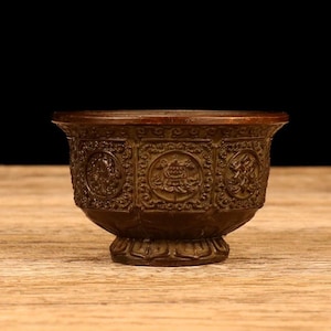 May include: A bronze-colored, ornate bowl with a wide rim and a detailed, hexagonal body. The bowl features embossed patterns and symbols, resting on a decorative base. The bowl is likely used for decorative or ceremonial purposes.