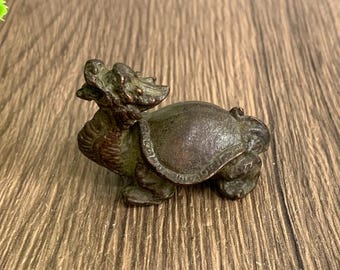 May include: A small, bronze-colored figurine of a mythical creature, a hybrid of a dragon and a turtle. The sculpture has intricate details, with a textured shell and a dragon-like head. The figurine is displayed on a wooden surface.