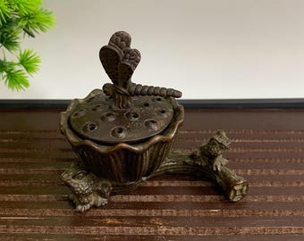 May include: Bronze incense burner in the shape of a lotus flower, with a cobra-like figure on top. The burner rests on a branch-like base, with a frog-like figure at the base. The color is a dark bronze.