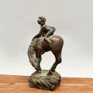 Puede incluir: Escultura de bronce que representa un mono montado a caballo. El mono está posado sobre la espalda del caballo, que se encuentra sobre una base texturizada. La escultura tiene un aspecto envejecido, con tonos marrones y verdes.