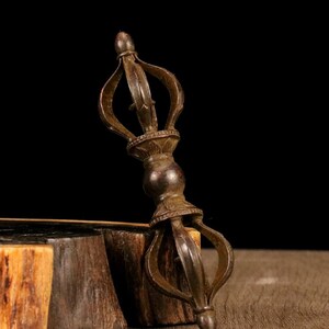 Könnte beinhalten: Ein bronzefarbener Vajra, ein rituelles Objekt mit symmetrischem Design. Das Objekt hat eine zentrale Kugel, verzierte Details und gebogene Zacken, die sich von oben und unten erstrecken. Es wird auf einer Holzoberfläche vor dunklem Hintergrund gezeigt.