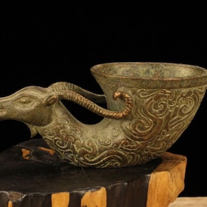 May include: An antique bronze cup with a ram's head design. The cup features intricate scrollwork and a curved handle. The bronze has a green patina, and the cup is displayed on a wooden base.