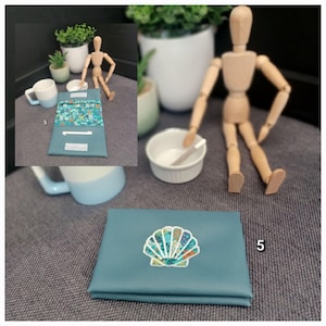 May include: Teal-colored wallet with a seashell design. The wallet is closed and has a stitched seashell design on the front. The interior of the wallet has a floral pattern. The number 5 is visible on the wallet.