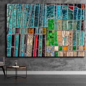 May include: Large abstract wall art featuring a mosaic of rectangular shapes in shades of teal, green, red, and gold. The artwork hangs on a textured gray wall, with a gray chair and potted plant in the foreground.