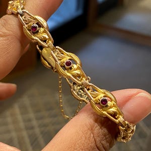 May include: A gold bracelet featuring a series of repeating ornate links with leaf-like motifs, each centered with a small, round, deep red gemstone. The metalwork displays a textured, polished finish, and a thin safety chain is attached to the clasp.