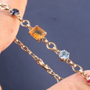 Pode incluir: Um bracelete de corrente de ouro com pedras preciosas coloridas. O bracelete apresenta uma pedra preciosa laranja de forma quadrada, uma pedra preciosa azul redonda e duas pedras preciosas vermelhas redondas.