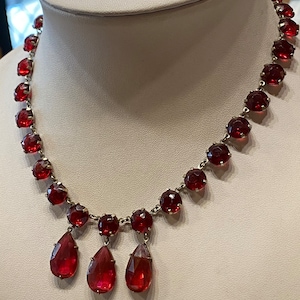 May include: A red gemstone necklace with three teardrop-shaped pendants. The necklace is made of silver and features round red gemstones.