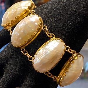 Può includere: Un braccialetto a catena d'oro con quattro grandi pietre ovali di madreperla. Le pietre sono incastonate in montature dorate con bordi a festoni.