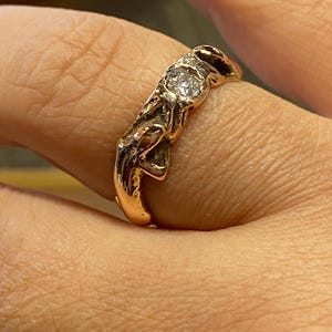 May include: A gold-colored ring with a textured, organic design. The ring features a small, round, clear gemstone set in the center. The band has an intricate, sculpted appearance, with a slightly uneven surface.