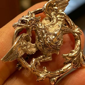 Pode incluir: Um broche de metal prateado com uma figura detalhada de um grifo com as asas abertas, posicionado dentro de uma moldura circular decorada com padrões ornamentais de folhas e vinhas. A criatura mítica é representada de perfil.