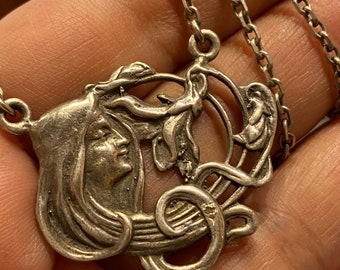 May include: A silver pendant necklace featuring an Art Nouveau design of a woman's face with flowing hair and a floral motif. The pendant is attached to a silver chain.