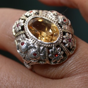 Puede incluir: Un anillo de plata con una piedra preciosa grande de color amarillo de forma ovalada rodeada de piedras preciosas rojas más pequeñas y diseños florales intrincados.