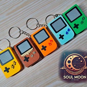 以下が含まれることがあります： 黄色、茶色、オレンジ、青、緑の5色のキーホルダーゲーム機コレクション。各キーホルダーには、シルバーのリングとチェーン、黒い十字と長方形の画面が付いています。Soul Moon 3D Designのロゴが右下にあります。
