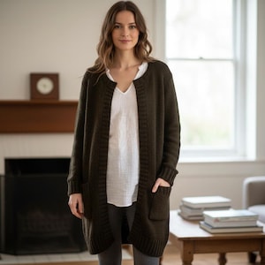 May include: A woman wearing a long, dark olive green cardigan sweater with two front pockets. The sweater is open in the front and worn over a white top. The woman is standing in a living room with a fireplace and a window.