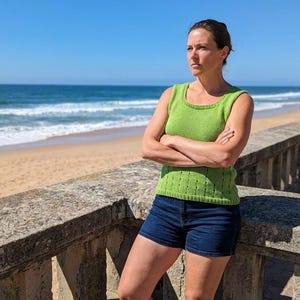 May include: A woman wearing a green knitted tank top and dark blue denim shorts stands with arms crossed. The top has a square neckline and a textured pattern. The background shows a beach, ocean, and clear blue sky.