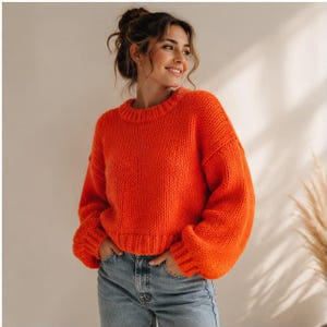 May include: A bright orange chunky knit sweater with a crew neckline, long sleeves, and ribbed cuffs. The sweater features a thick, textured yarn construction with a relaxed fit, paired with light blue denim jeans that have a classic button fly.