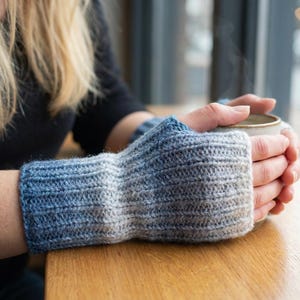 May include: Hand-knitted fingerless gloves in a gradient of blue and gray. The wrist cuffs are a deep blue, fading to a lighter gray at the fingers. The gloves are made of a soft, textured yarn, perfect for keeping hands warm.