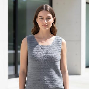 May include: A gray crochet tank top with a scoop neckline. The sleeveless top is made of a textured, knitted material. The model is wearing the top in front of a modern building.