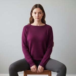 Op de afbeelding: Een persoon draagt een donkerpaarse trui met een ronde hals en lange mouwen, gecombineerd met een grijze legging. De persoon zit op een houten stoel tegen een lichtgrijze achtergrond.