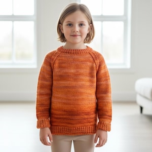 May include: An orange knitted sweater with a crew neck and long sleeves. The sweater has a ribbed hem and cuffs. The child is wearing beige trousers. The background is a bright room with windows and a sofa.