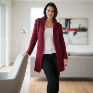 May include: A burgundy knit cardigan with a shawl collar. The long-sleeved sweater is worn over a white top and black pants. The woman is standing near a gray chair in a well-lit room.