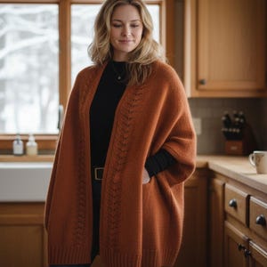 May include: A rust-colored knit cardigan with a decorative openwork design. The cardigan is worn over a black top and jeans. The woman is standing in a kitchen with wooden cabinets and a window.