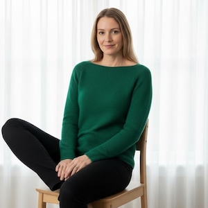 May include: A woman wearing a green long-sleeve sweater and black pants sits on a wooden chair. The sweater has a boat neck design. The background is a white curtain.