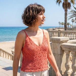 Puede incluir: Un camisola de punto color coral con cuello en V y un patrón texturizado. La parte superior tiene tirantes finos y un dobladillo festoneado. La imagen está tomada al aire libre con una playa y palmeras al fondo.