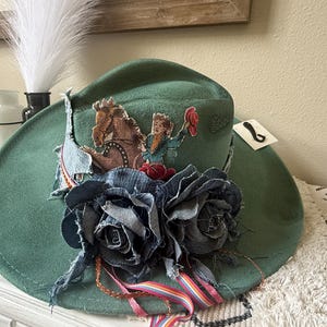 May include: A green cowboy hat with a wide brim, decorated with a denim flower and a vintage-style image of a cowboy on a horse. The hat also features a small tag and colorful ribbons.