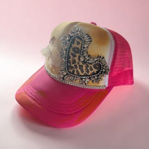 May include: A pink trucker hat with a leopard print heart design embellished with rhinestones. The hat features a pink brim, a mesh back, and a tan and white front panel. A small cluster of white feathers is attached to the side.