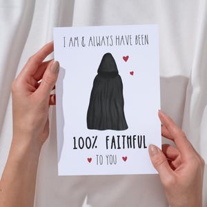 May include: A white greeting card with the text "I AM & ALWAYS HAVE BEEN" and "100% FAITHFUL TO YOU." A black hooded cloak illustration is centered on the card, with three red hearts. The card is held by two hands.