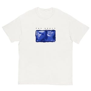 以下が含まれることがあります： 2人の顔の青いグラフィックが描かれた白いTシャツ。上には「far apart」という文字が書かれている。