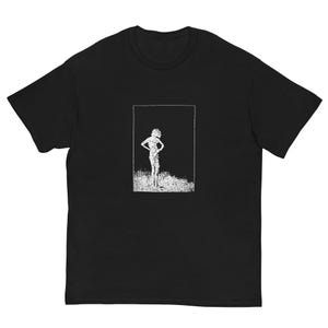 May include: Black t-shirt featuring a white graphic of a person standing with hands on hips, framed by a white rectangle. The graphic has a textured, grainy appearance. The shirt is a casual wear item.